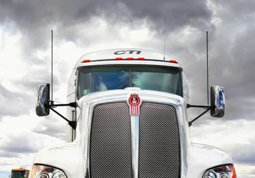 Close-up of the front of a large white semi-truck with a grille, windshield, and side mirrors, under a cloudy sky.