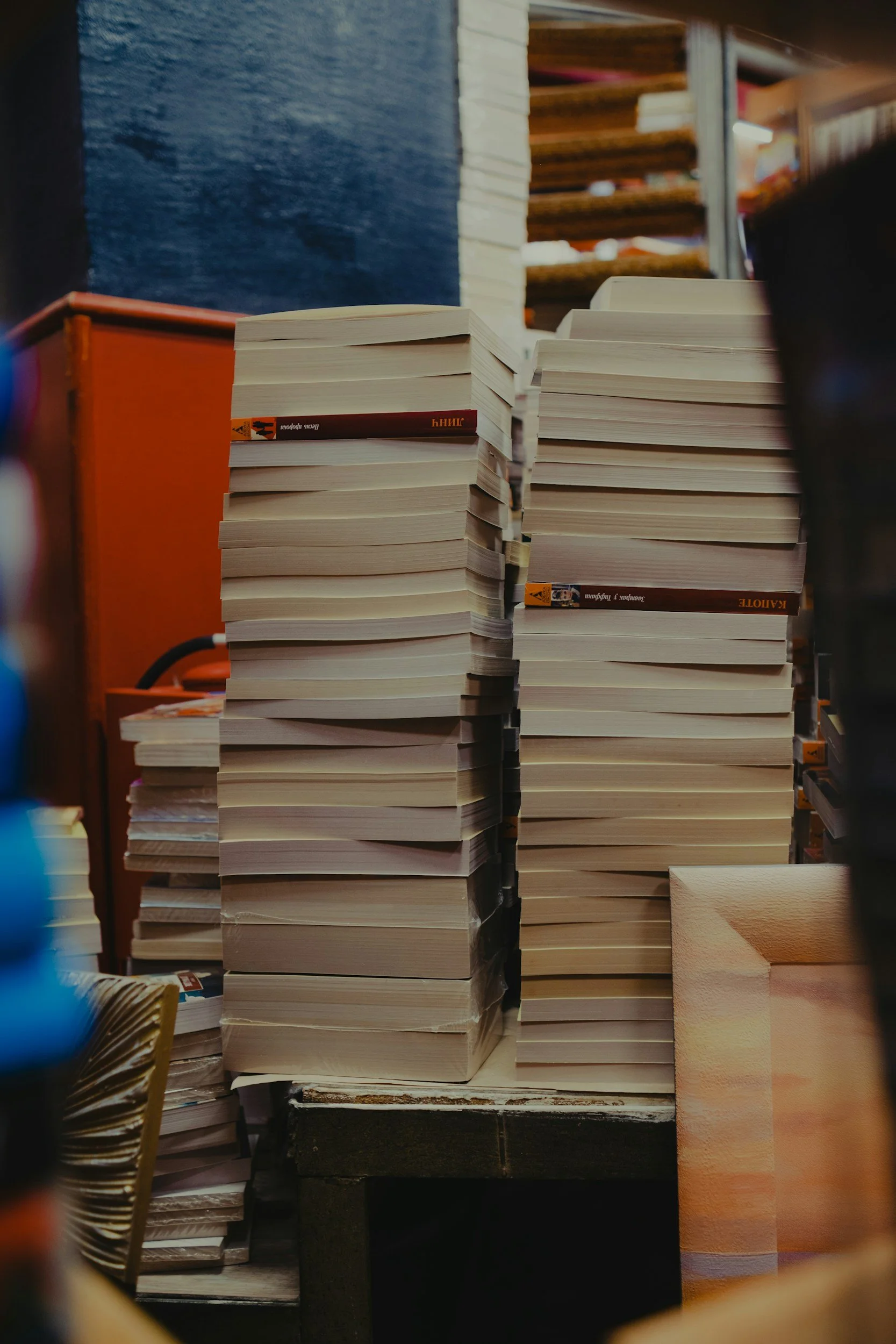 Photo of a stack of books on a table, with a blue wall and wooden shelves filled with books in the background.