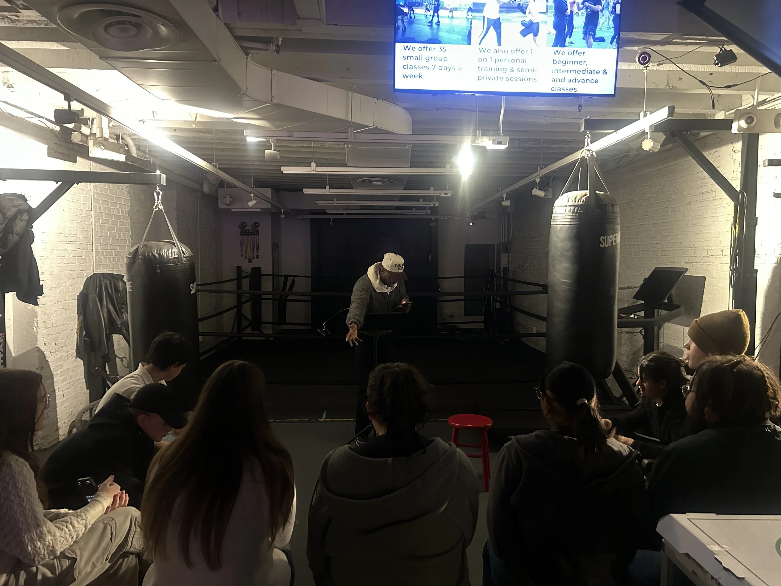 A group of people seated in a boxing gym watching a boxing match or seminar with boxing bags hanging in the foreground and a large digital screen displaying information above.