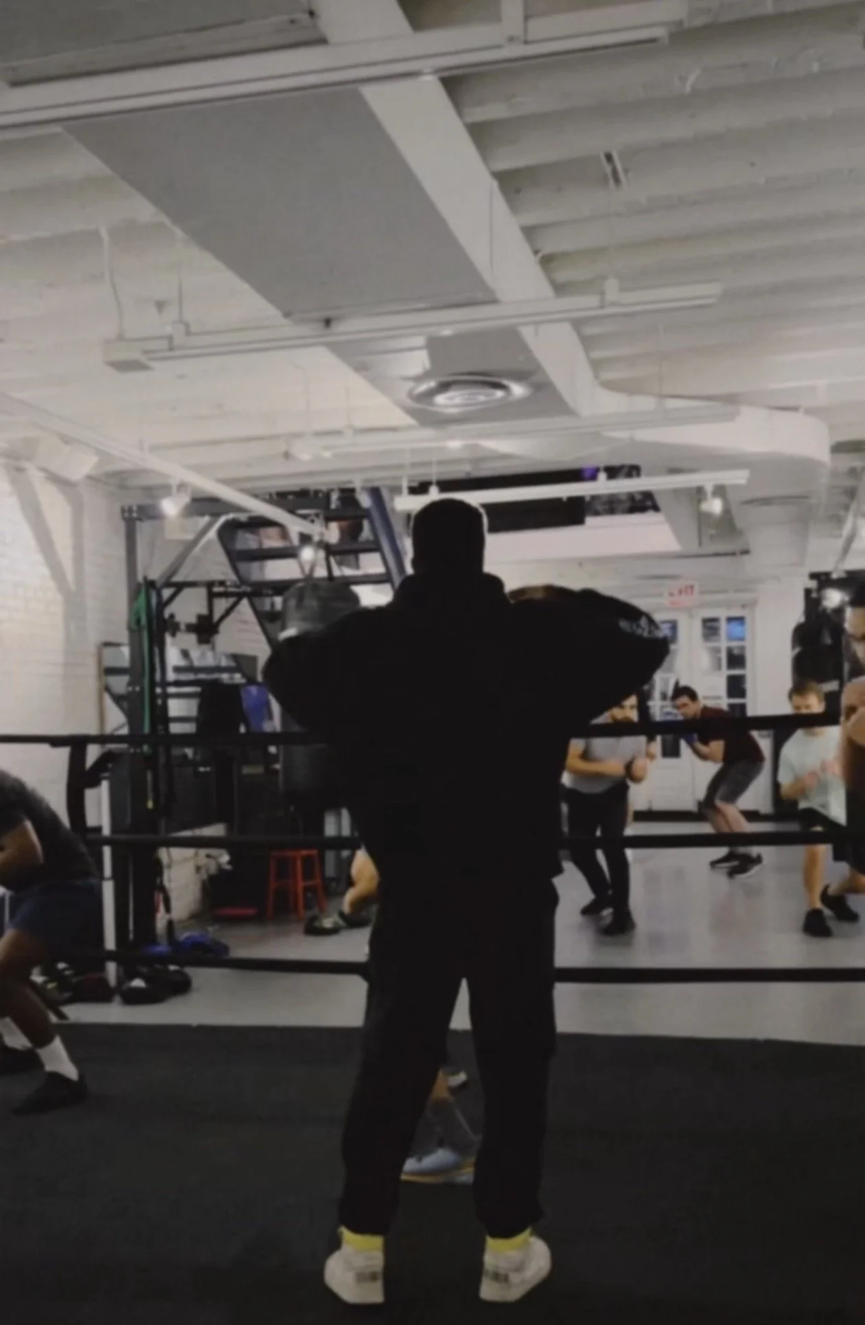 People training in an indoor boxing gym with a boxer silhouetted in the foreground and a heavy bag in the background.