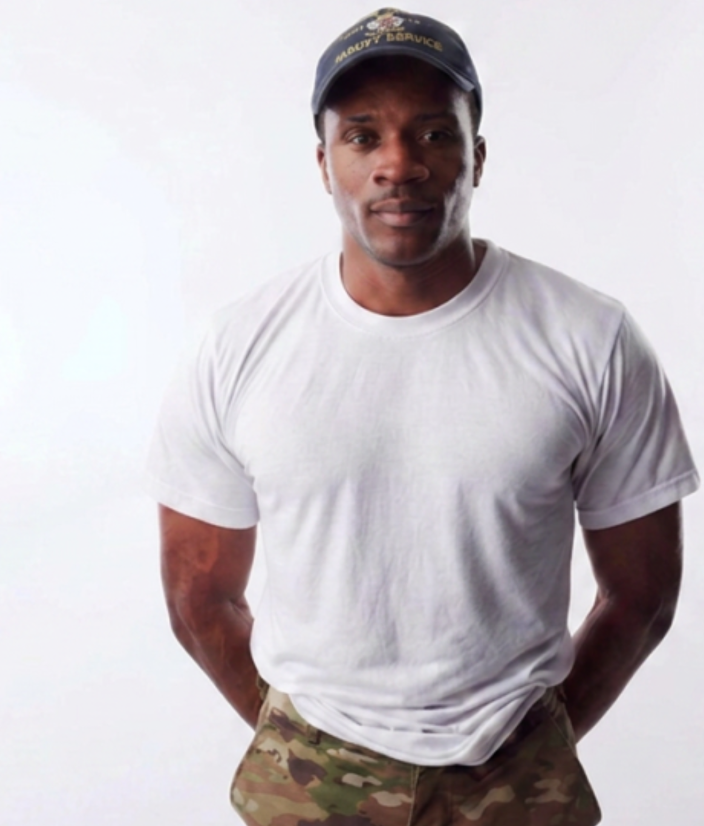 Coach Adres in a white t-shirt, army patterned pants, and a navy baseball cap standing with his arms behind his back in a strong athletic stance