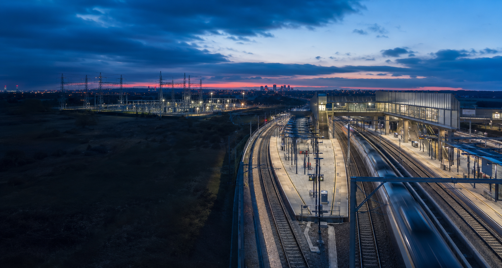 A modern train station at dusk with multiple train tracks, platforms, and a train arriving or departing, under a sky with dark clouds and city lights in the background.