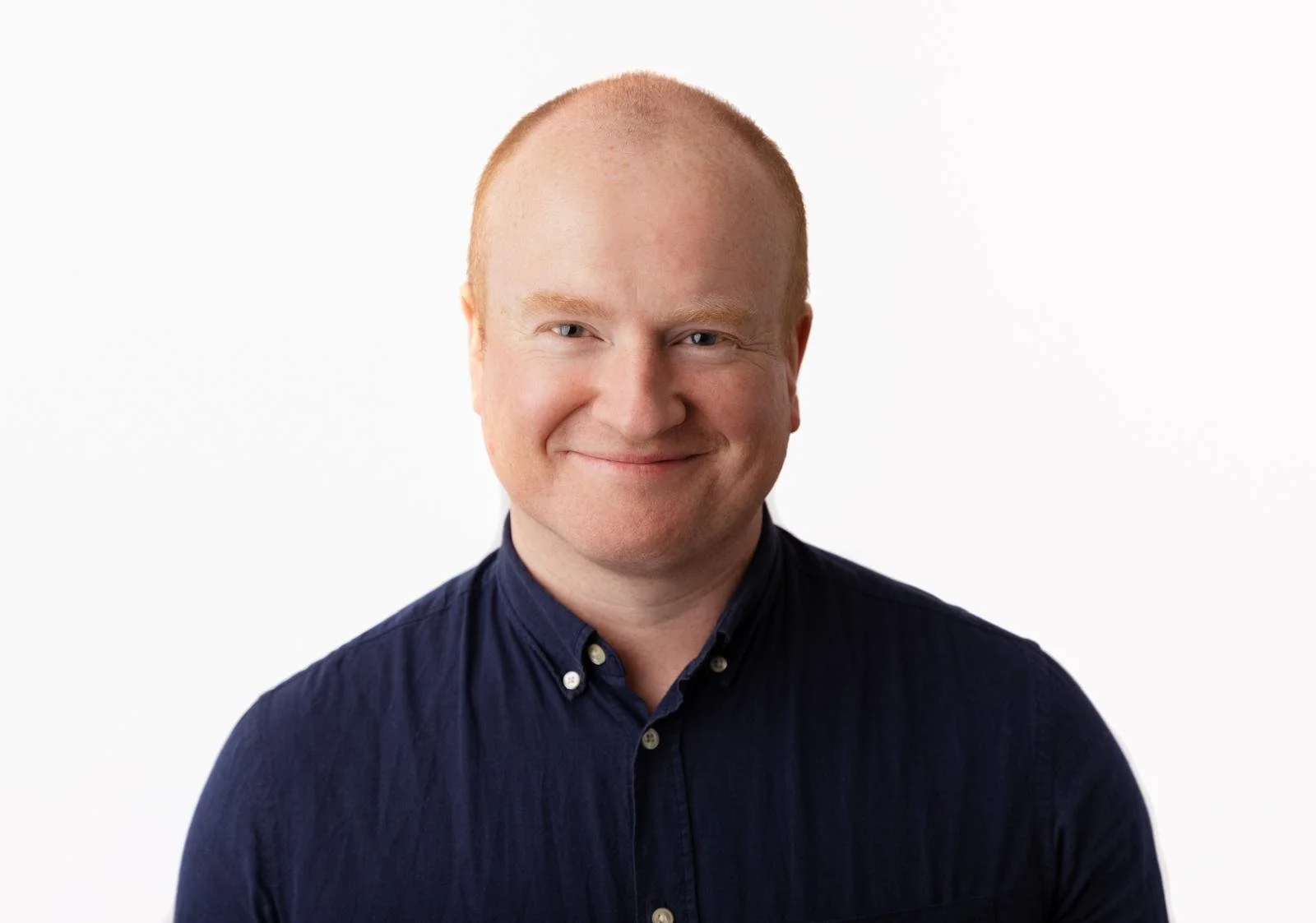 Portrait of a smiling man with short reddish hair, wearing a dark blue button-up shirt, against a plain white background.