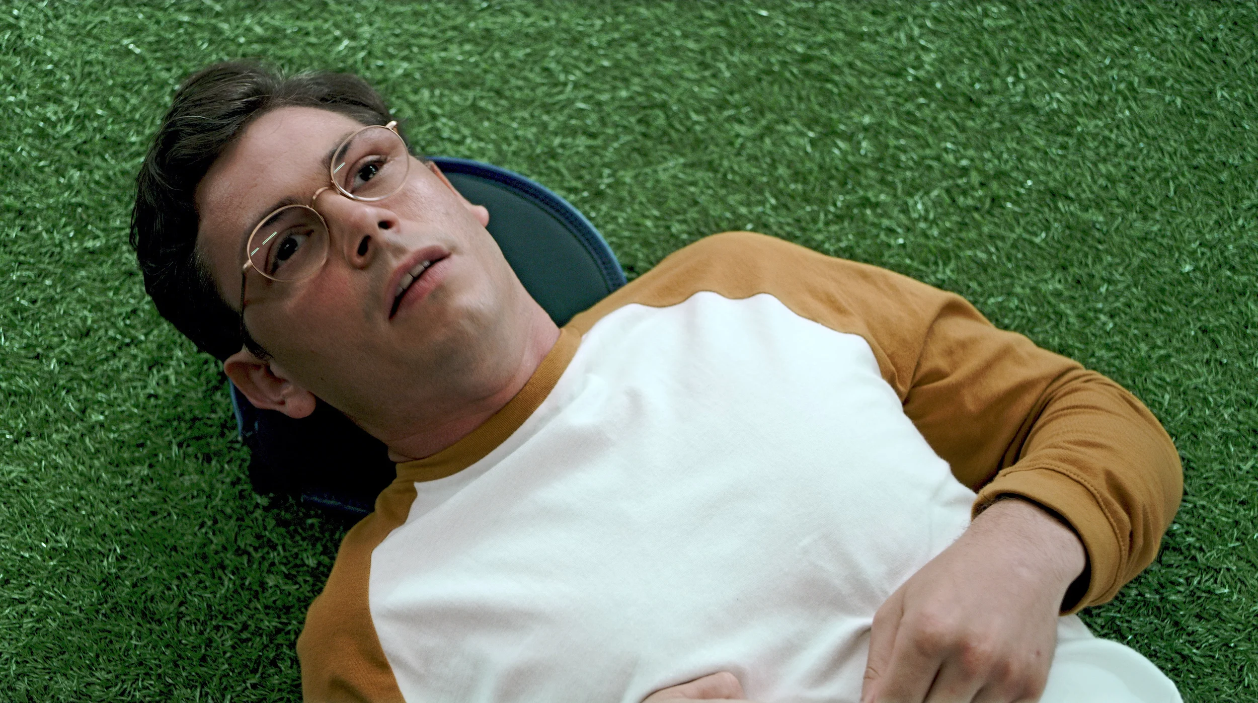 Ryan O'Connell wearing glasses and a yellow raglan shirt lying down on artificial green grass looking up.