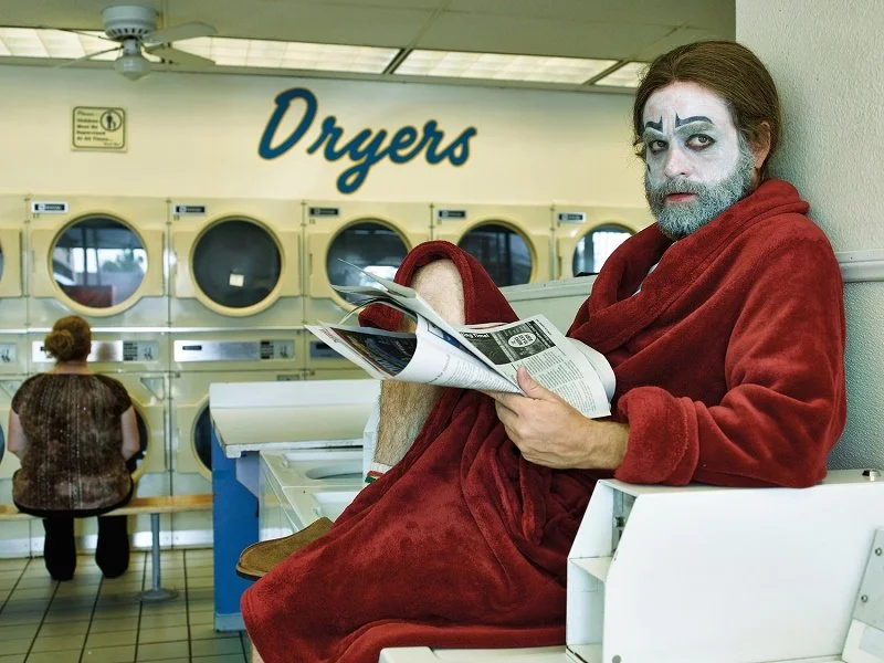 Zach Galifianakis in clown makeup and a plush red bathrobe sitting on a counter in a laundromat reading a newspaper.