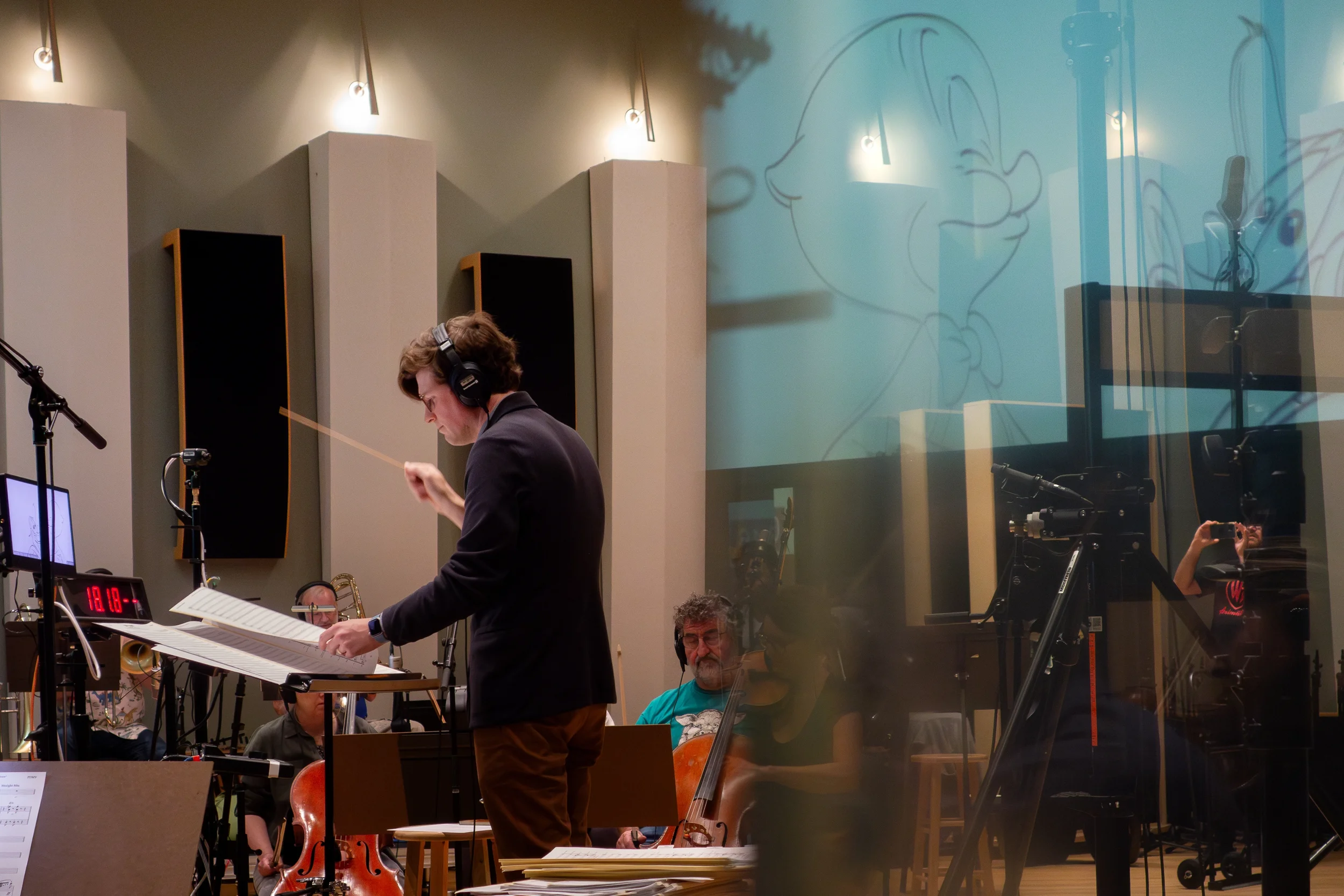Joshua Moshier raising his baton in a recording studio with a sketch outline of Porky Pig reflected on the control room glass.