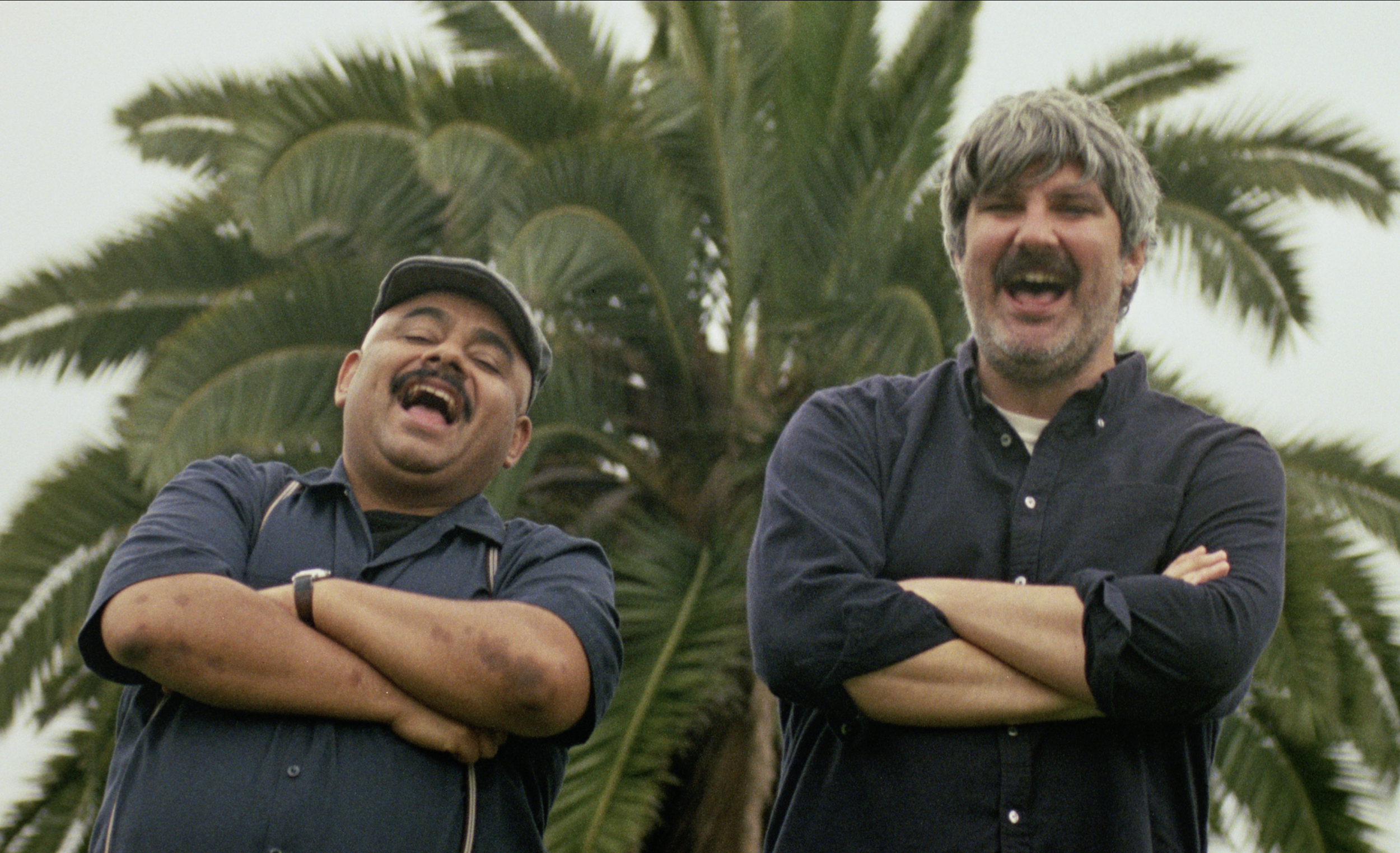 Two men standing side-by-side with arms crossed, laughing outdoors in front of palm trees in The Weird Guy.