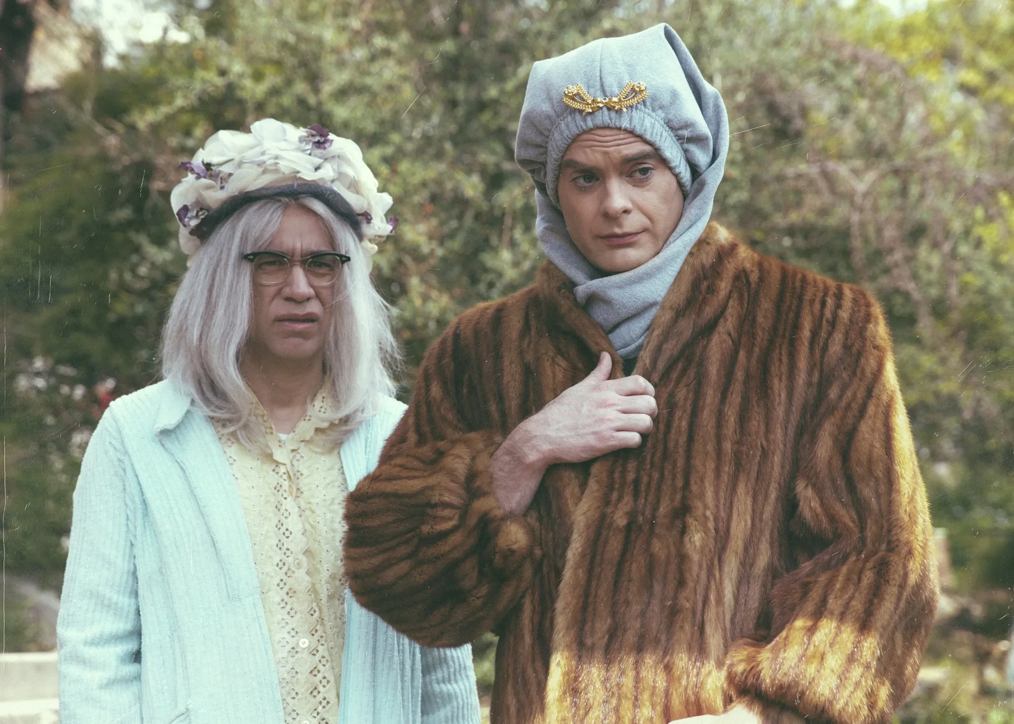 Fred Armisen in a gray wig and sunhat standing beside Bill Hader in a headscarf and fur coat outdoors.