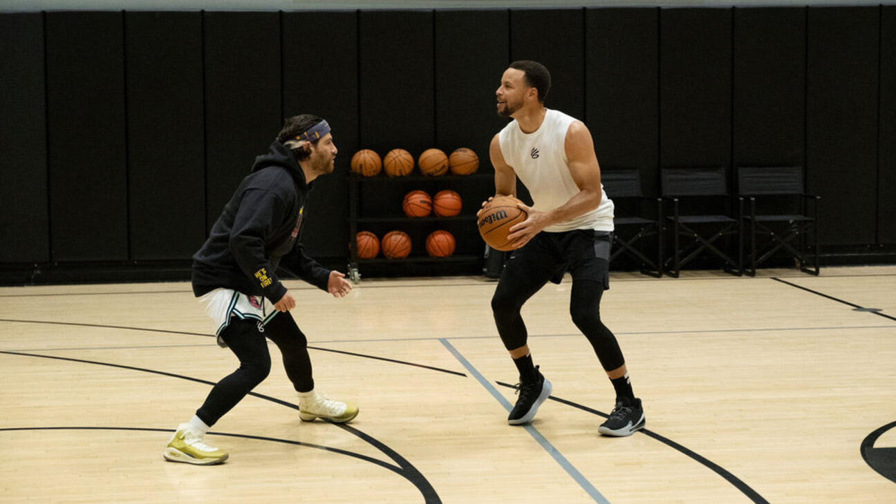 Adam Pally in a headband playing defense against Steph Curry holding a basketball on an indoor gym court.