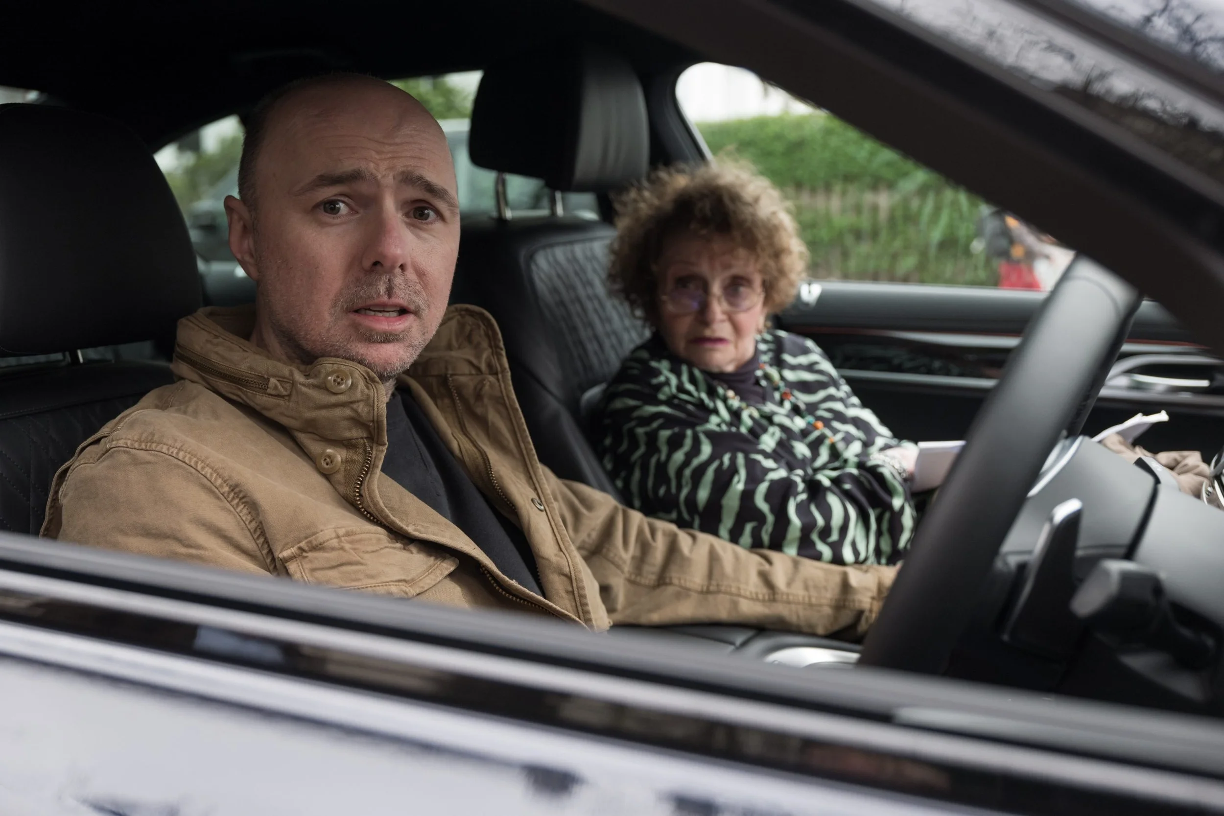 Karl Pilkington looking stressed in the driver's seat of a car next to an elderly woman.