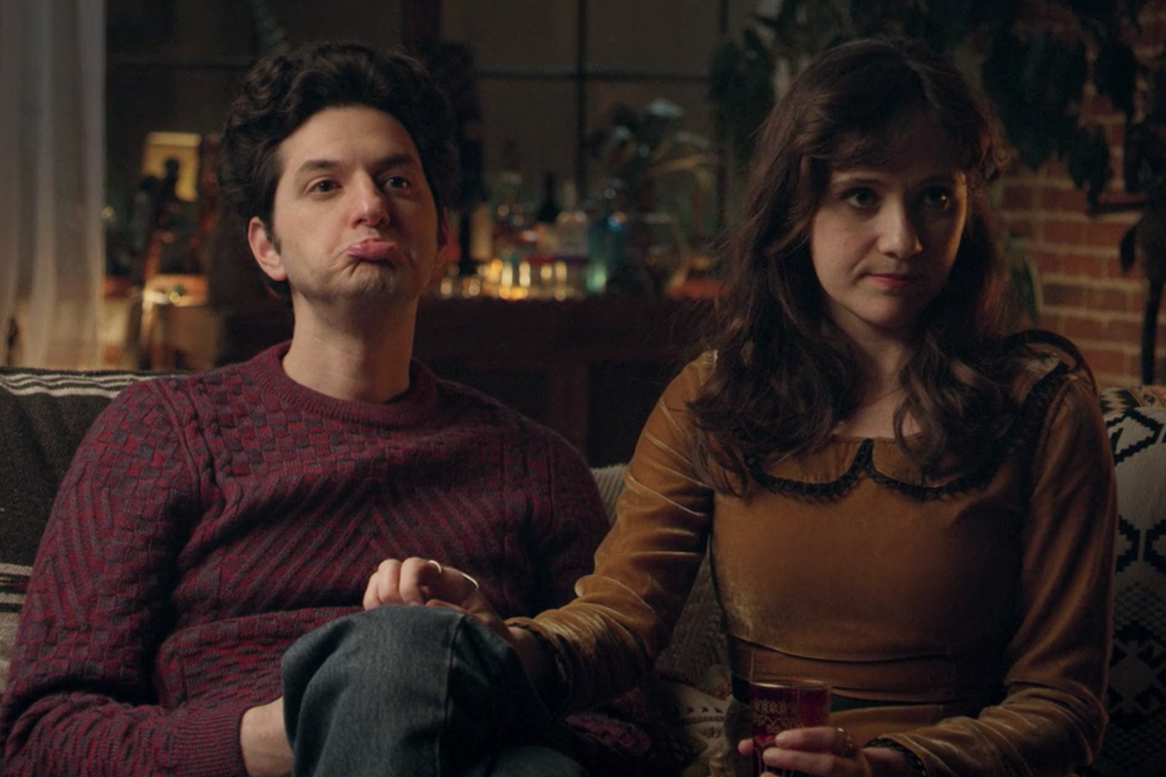 Ben Schwartz puffing his cheeks while sitting next to Noël Wells on a couch in Happy Anniversary.