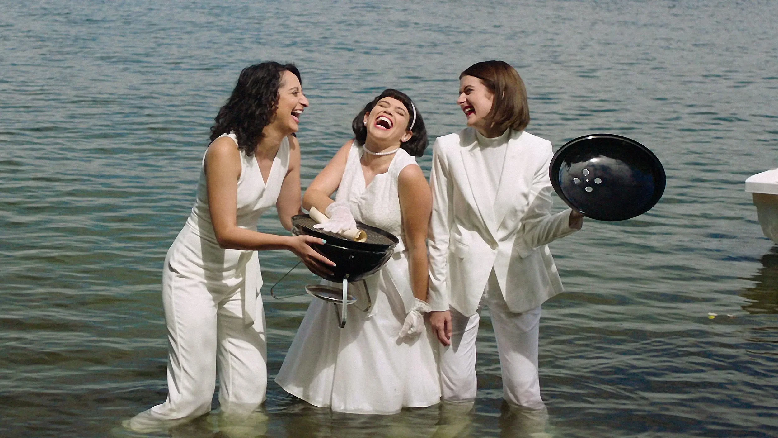 Sandy Honig, Mitra Jouhari, and Alyssa Stonoha wading waist-deep in water holding a barbecue grill in Three Busy Debras.