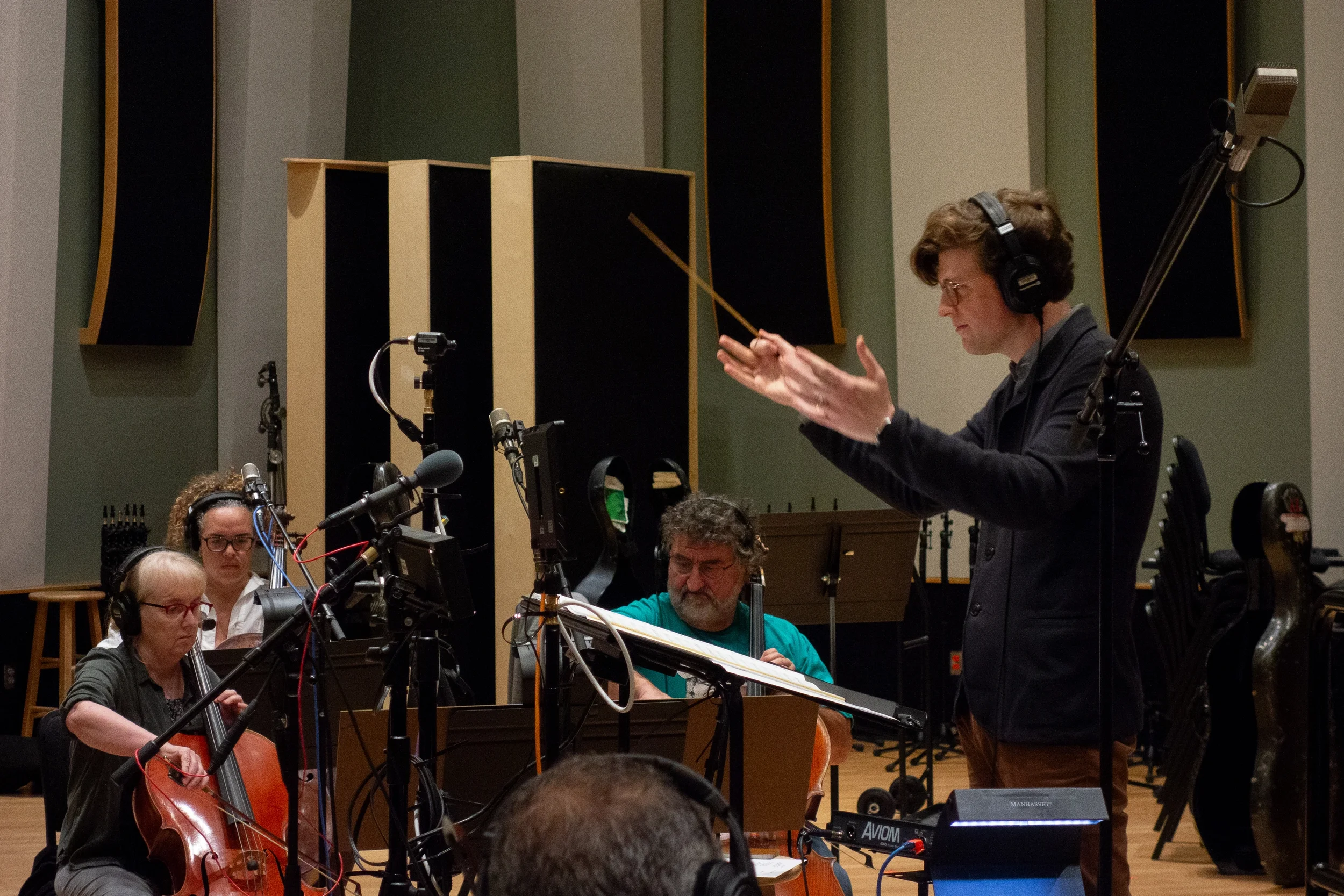 Joshua Moshier wearing headphones raising his hands and baton while leading a string ensemble in a sound studio.