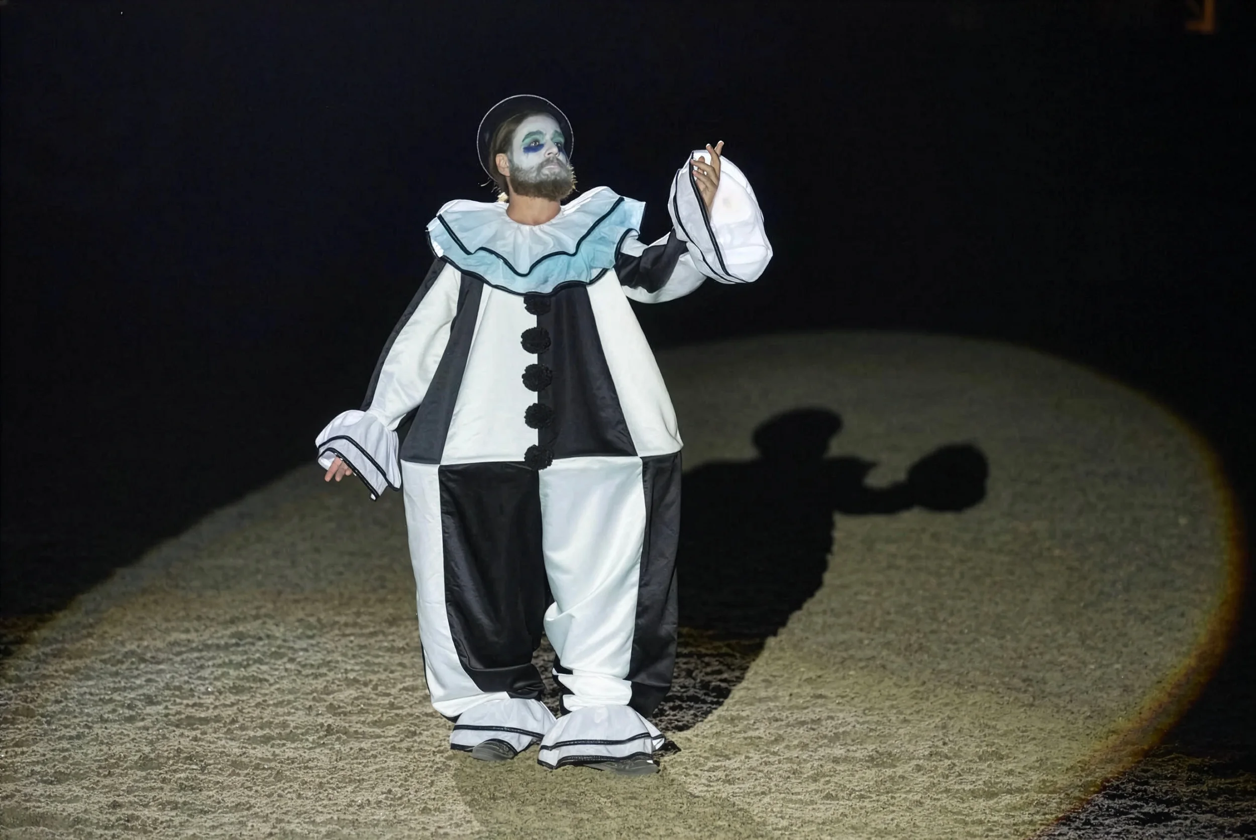 Zach Galifianakis performing in a black-and-white ruffled Pierrot clown costume under a spotlight.