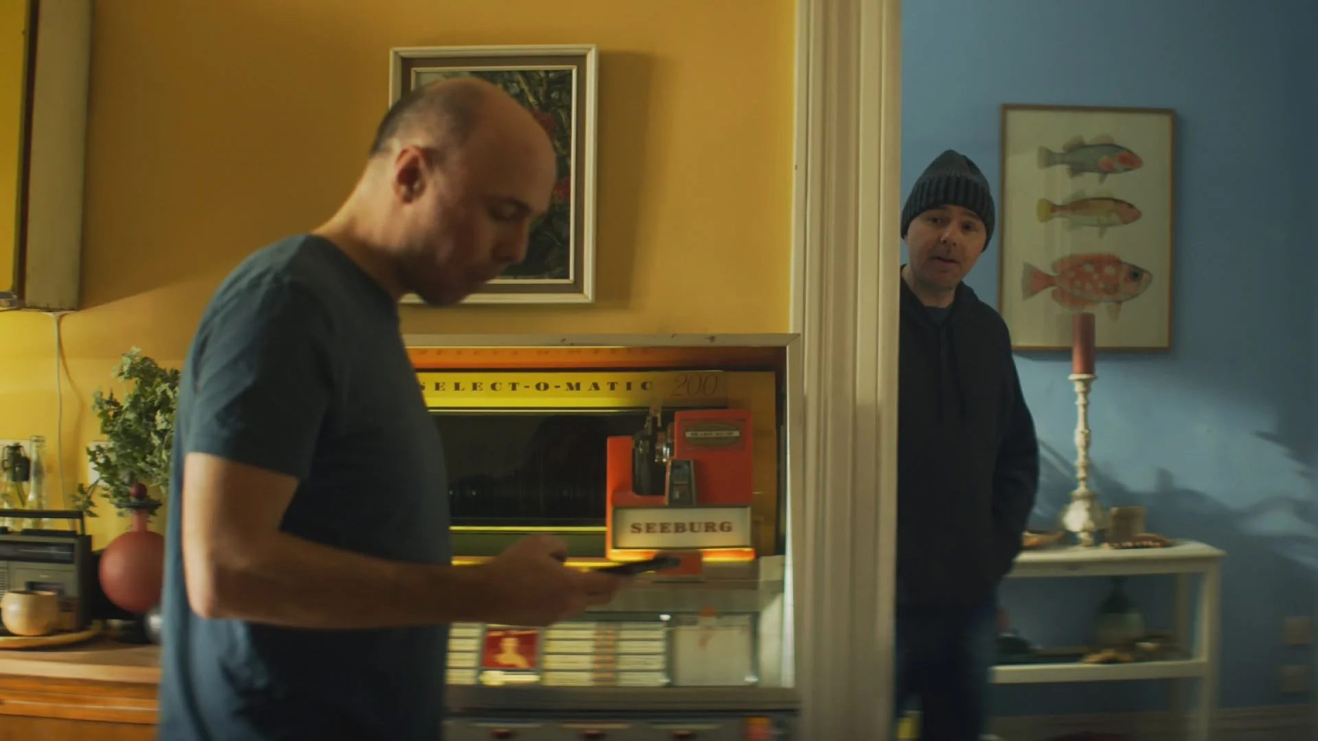 Karl Pilkington using his phone near a jukebox while his beanie-wearing inner voice peeks around a door frame.