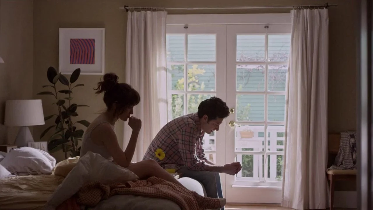 Noël Wells sitting on a bed while Ben Schwartz sits on a chair nearby in Happy Anniversary.