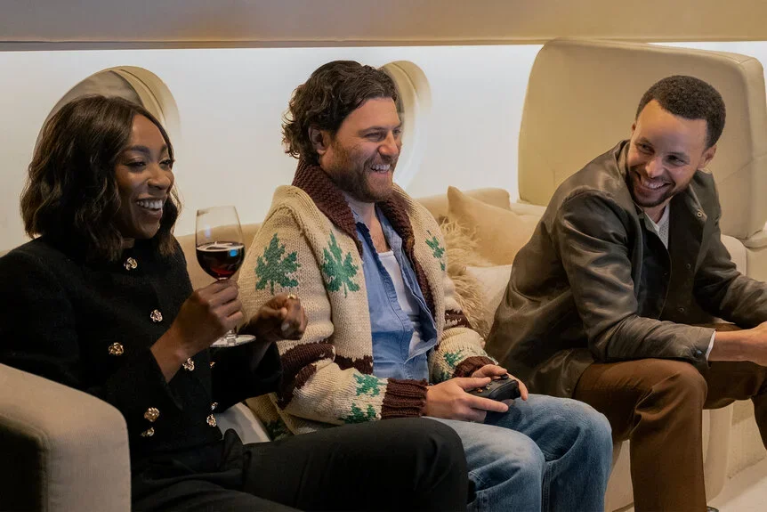 Ego Nwodim holding a glass of wine sitting beside Adam Pally and Steph Curry laughing inside a private jet.