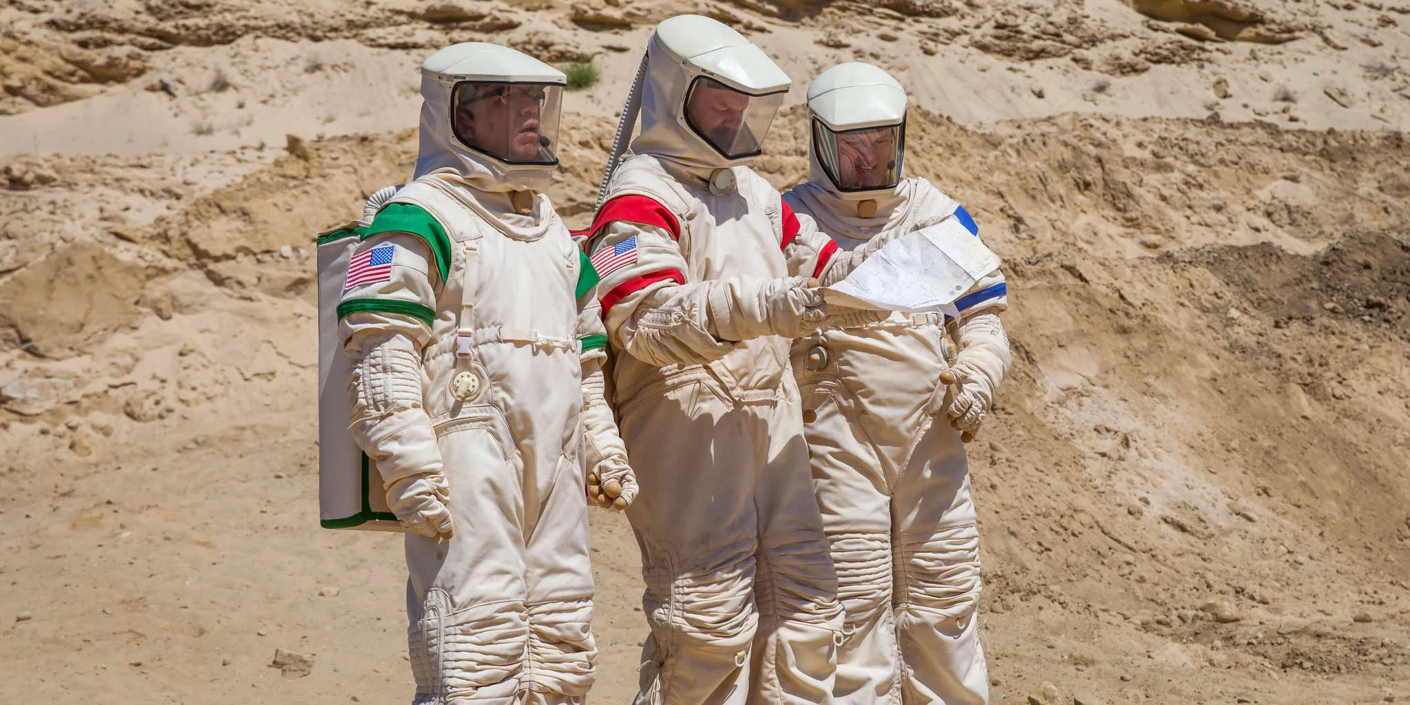 John C. Reilly, Tim Heidecker, and Fred Armisen wearing full white spacesuits in a desert training environment in Moonbase 8.