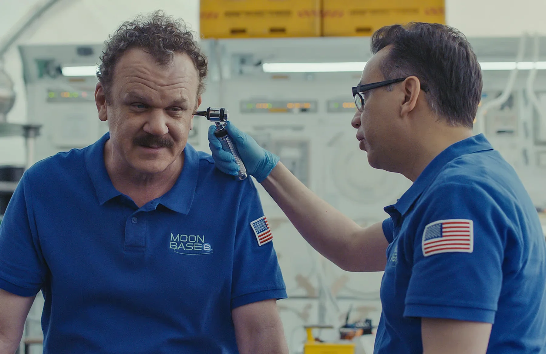 Fred Armisen examining John C. Reilly's ear with a medical tool inside the moonbase habitat in Moonbase 8.