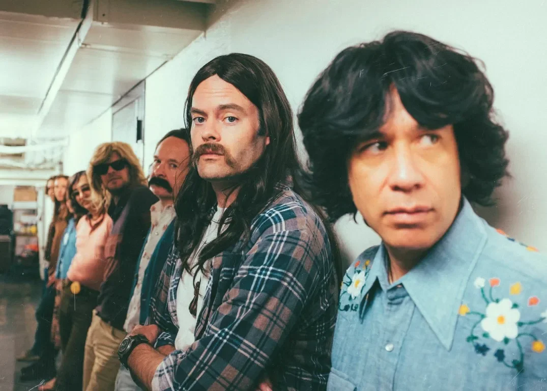 Bill Hader with long hair and a mustache standing next to Fred Armisen in an embroidered denim shirt with their band.