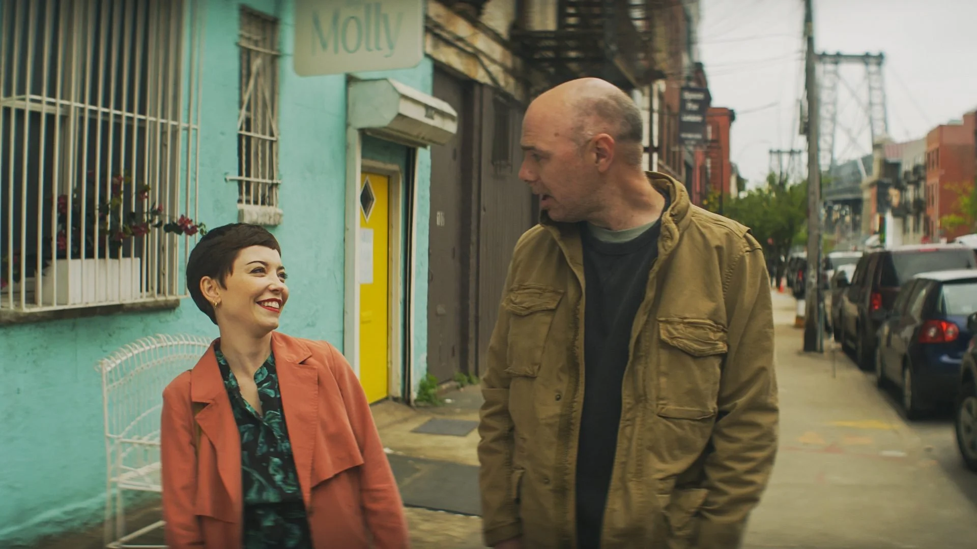 Karl Pilkington walking alongside a smiling woman in a pink coat outside a shop.