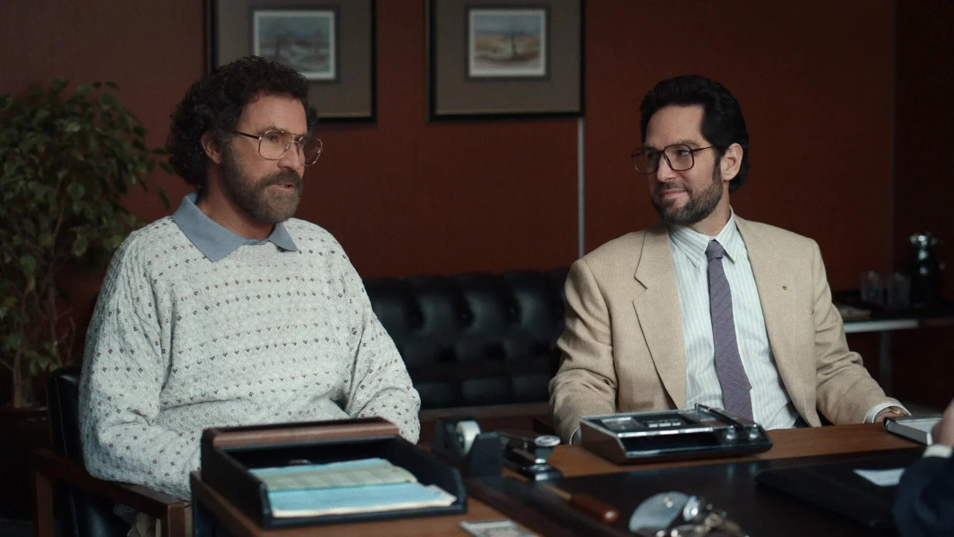 Will Ferrell in a patterned sweater and Paul Rudd in a beige suit sitting side-by-side at a desk during a meeting.