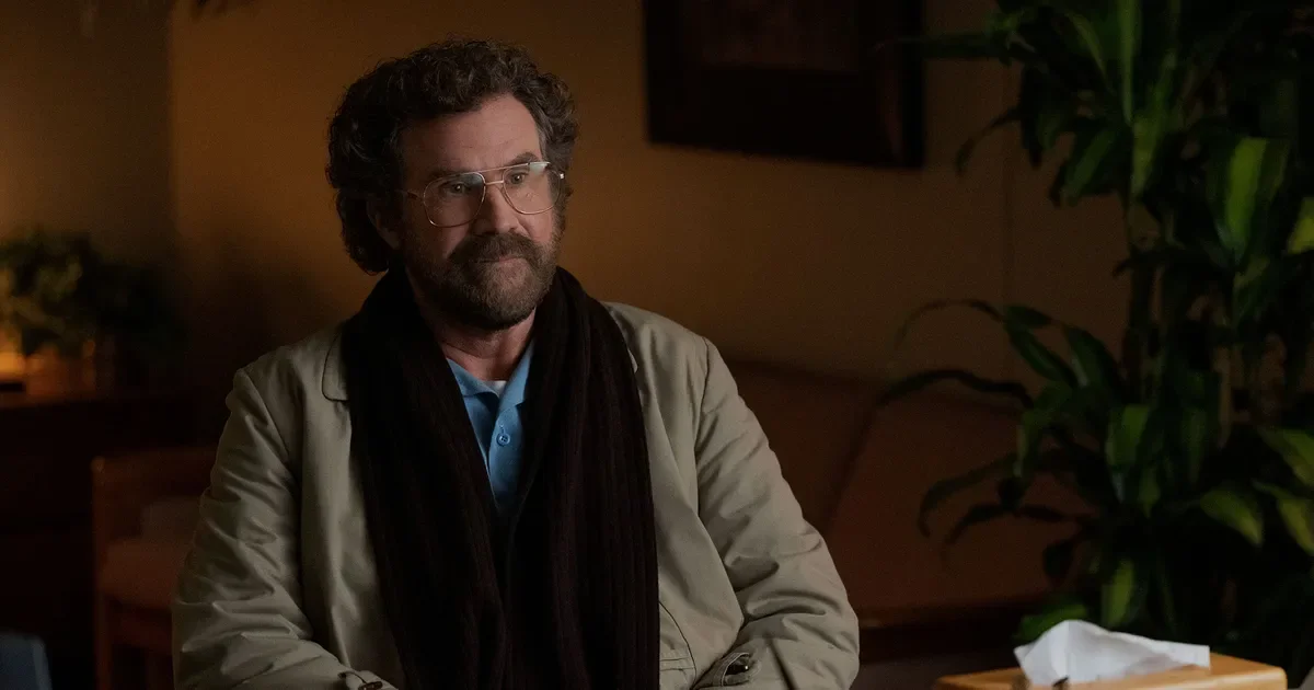 Close-up of Will Ferrell wearing a brown scarf, coat, and glasses, listening intently.