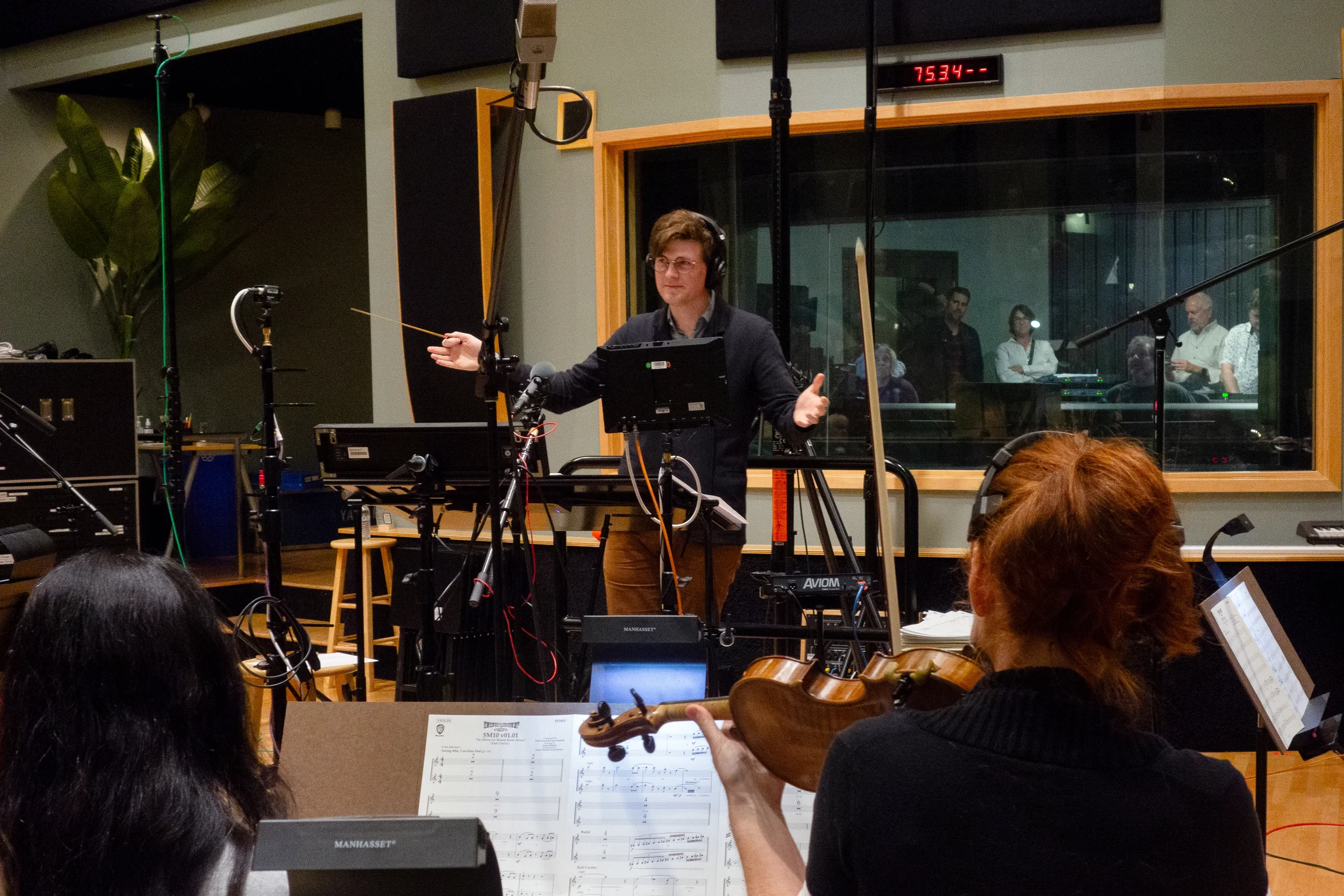 Joshua Moshier with headphones gesturing to musicians during a studio scoring session, seen over a violinist's shoulder.