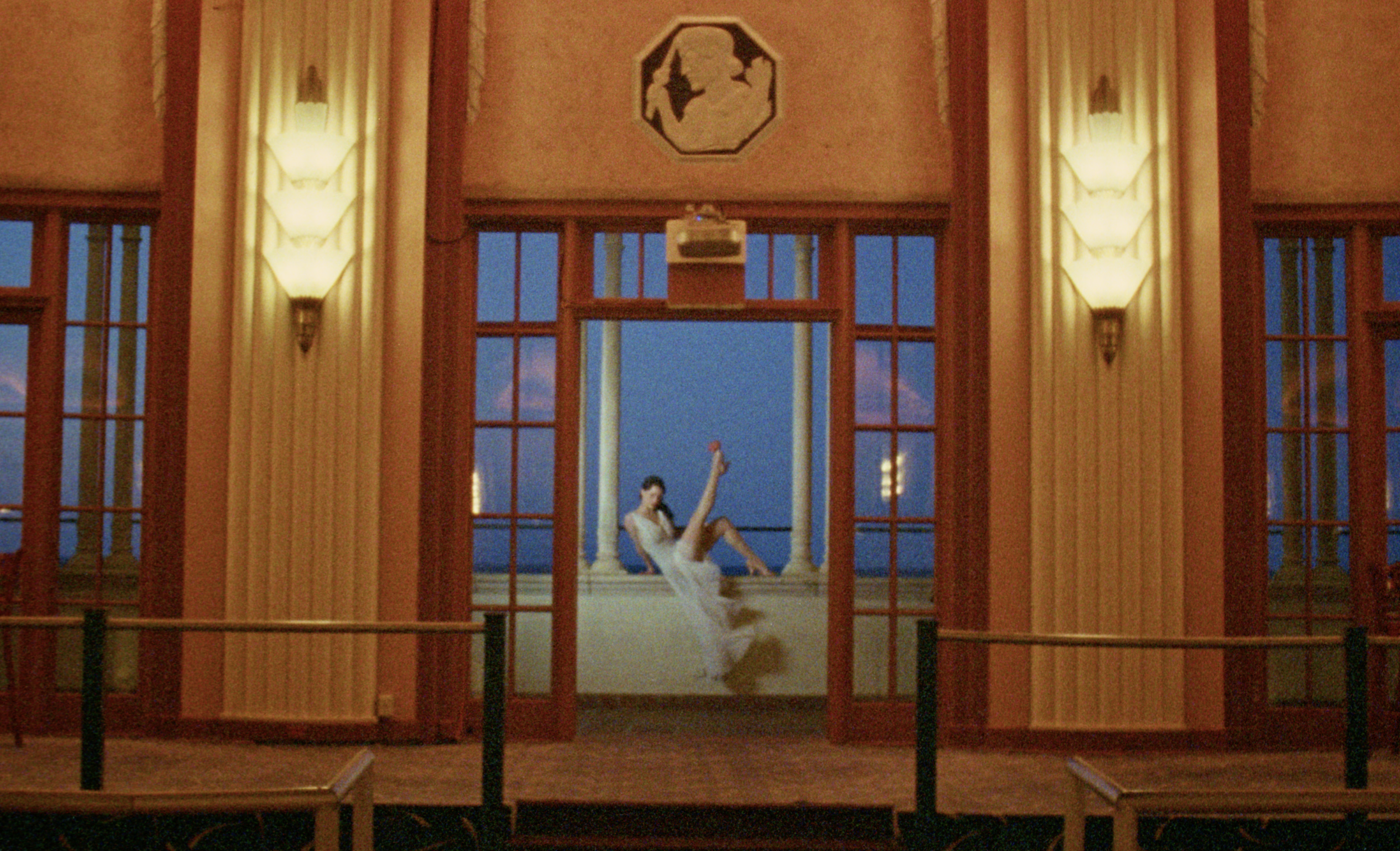 A woman sitting on a high window ledge wearing red heels inside an Art Deco corridor in The Weird Guy.