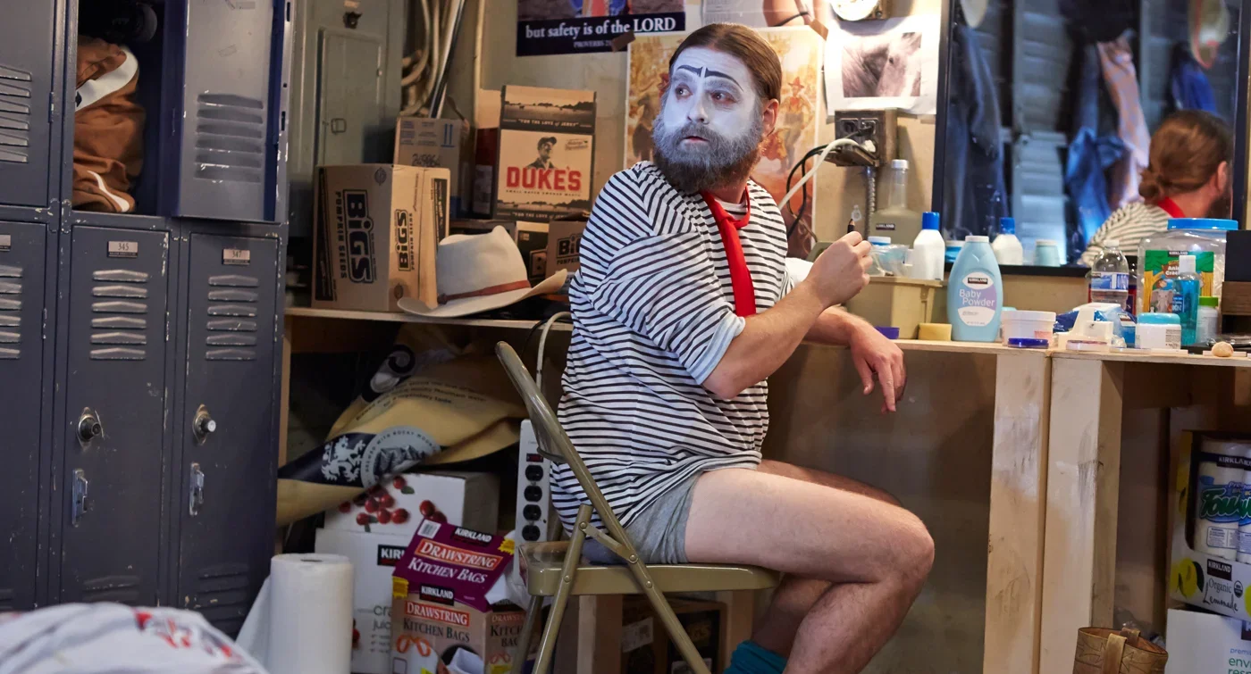 Zach Galifianakis wearing clown face paint, a striped shirt, and red scarf while sitting in a cluttered dressing room.