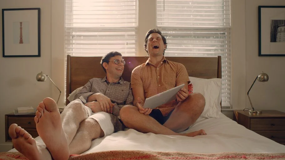 Two men sitting together on a bed laughing while looking at a tablet.