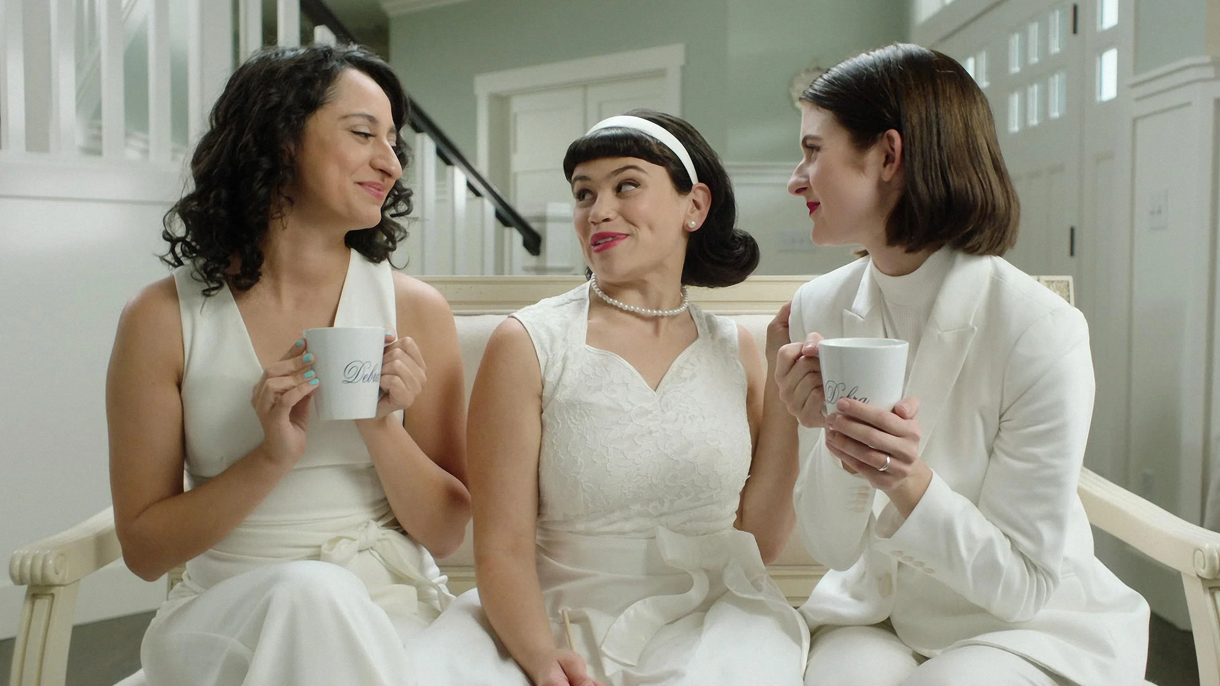 Sandy Honig, Mitra Jouhari, and Alyssa Stonoha dressed in all-white outfits sitting on a sofa with coffee mugs in Three Busy Debras.