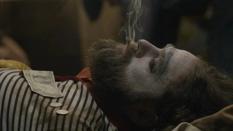 Close-up profile of Zach Galifianakis lying down smoking a cigarette with a dollar bill and coins on his chest.