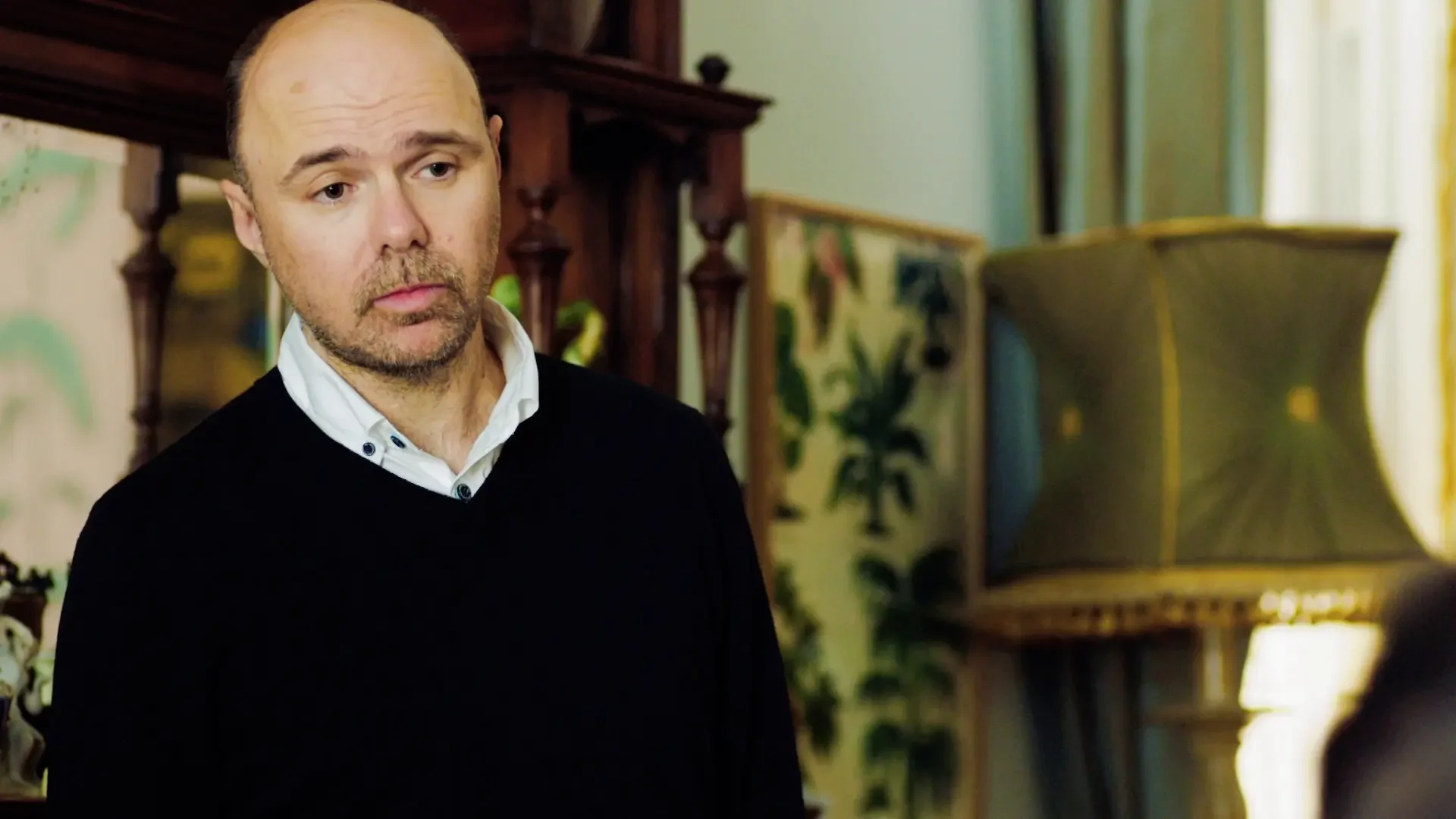 Close-up portrait of Karl Pilkington looking unamused in a black sweater and white collared shirt.