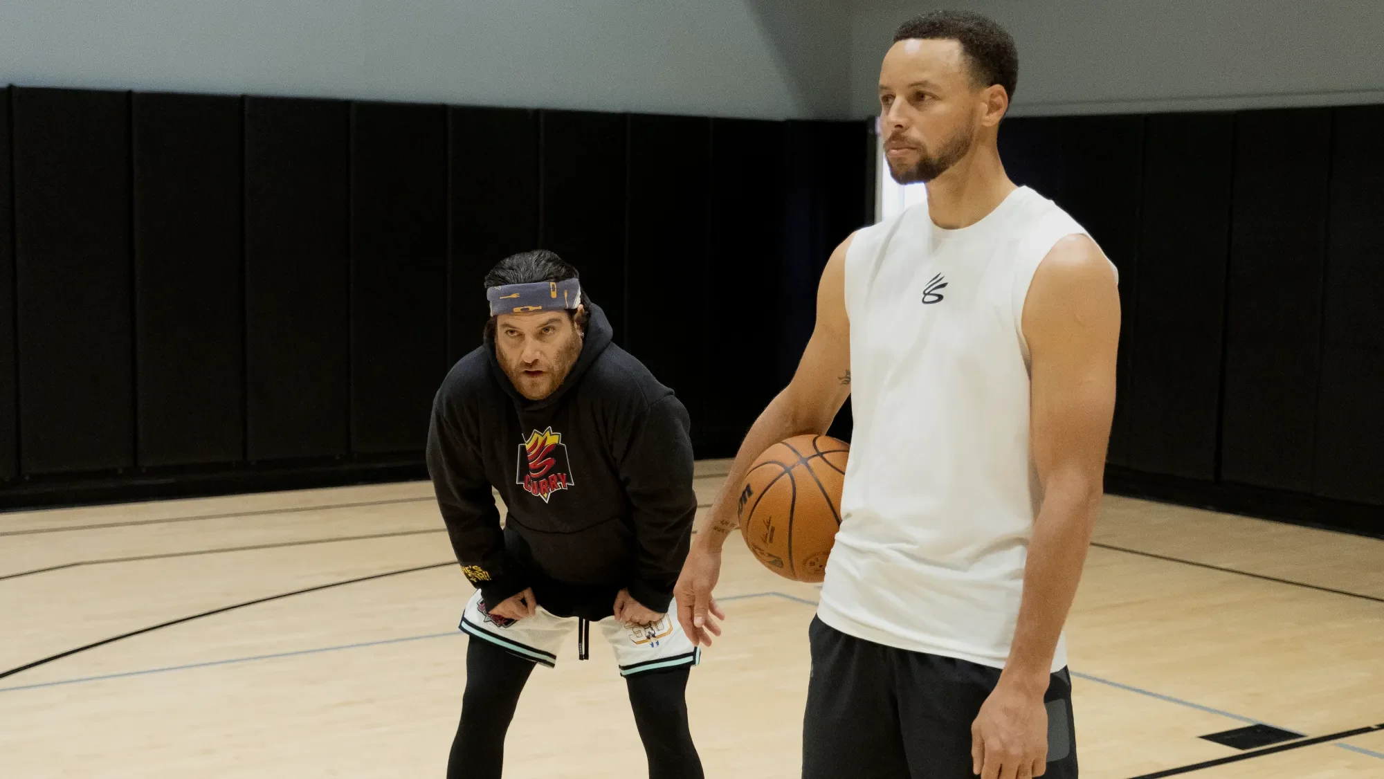 Adam Pally crouching behind Steph Curry holding a basketball.