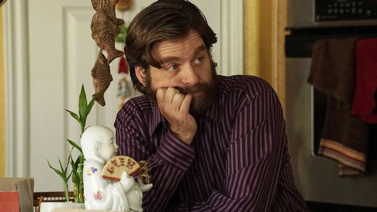 Zach Galifianakis in a purple striped shirt resting his chin on his hand with a thoughtful expression at a table with a small Buddha statue.