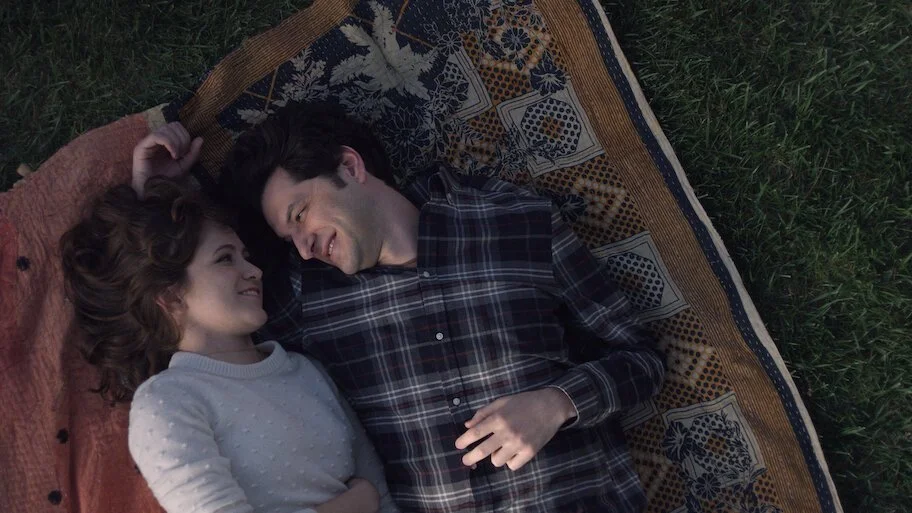 oël Wells and Ben Schwartz lying on a patterned blanket on the grass looking at each other.
