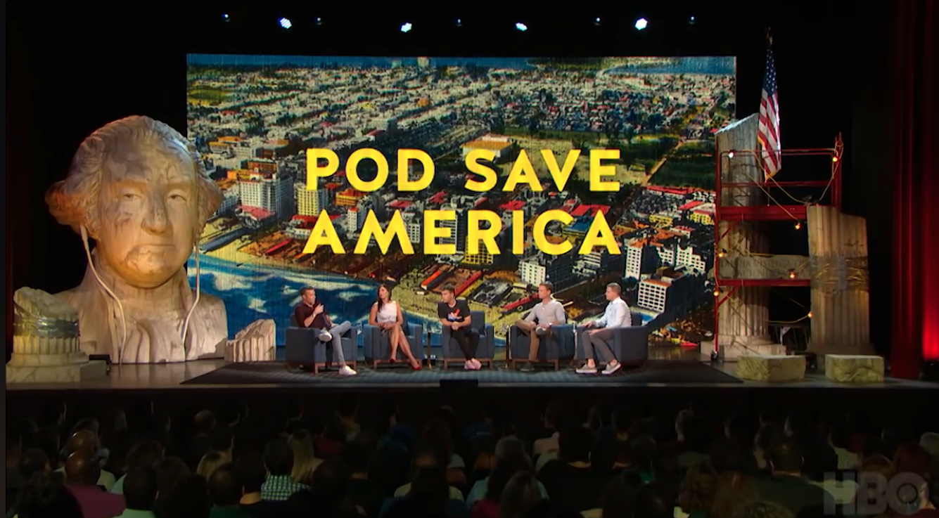 The hosts of Pod Save America seated on stage in front of a large backdrop screen and a George Washington head sculpture during a live recording.