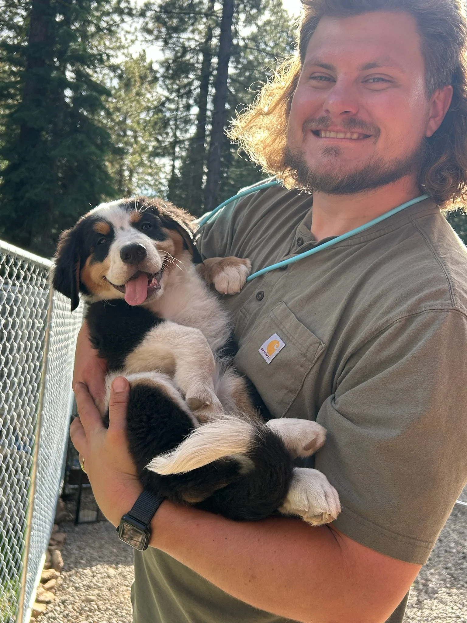 A man with long curly hair and a beard smiling while holding a tricolor puppy with black, white, and tan markings outdoors with trees in the background.