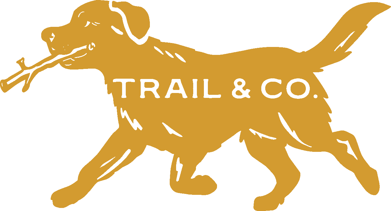 A gold-colored silhouette of a dog carrying a stick in its mouth, with the text 'TRAIL & CO.' inside the body of the dog.