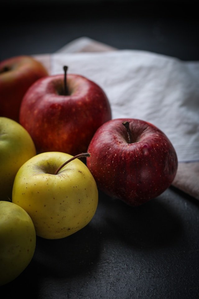 A close-up of several apples, with a mix of red and yellow varieties, on a dark surface with a crumpled white cloth in the background.
