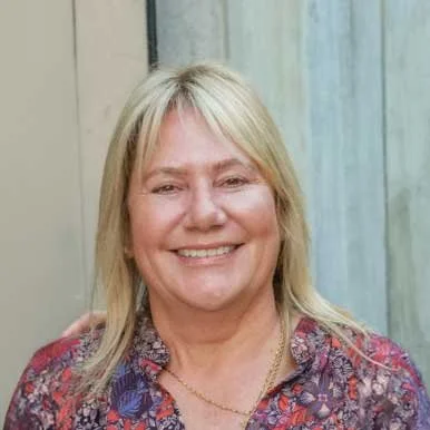 A smiling middle-aged woman with blonde hair wearing a patterned blouse and a gold necklace, standing in front of a wooden background.