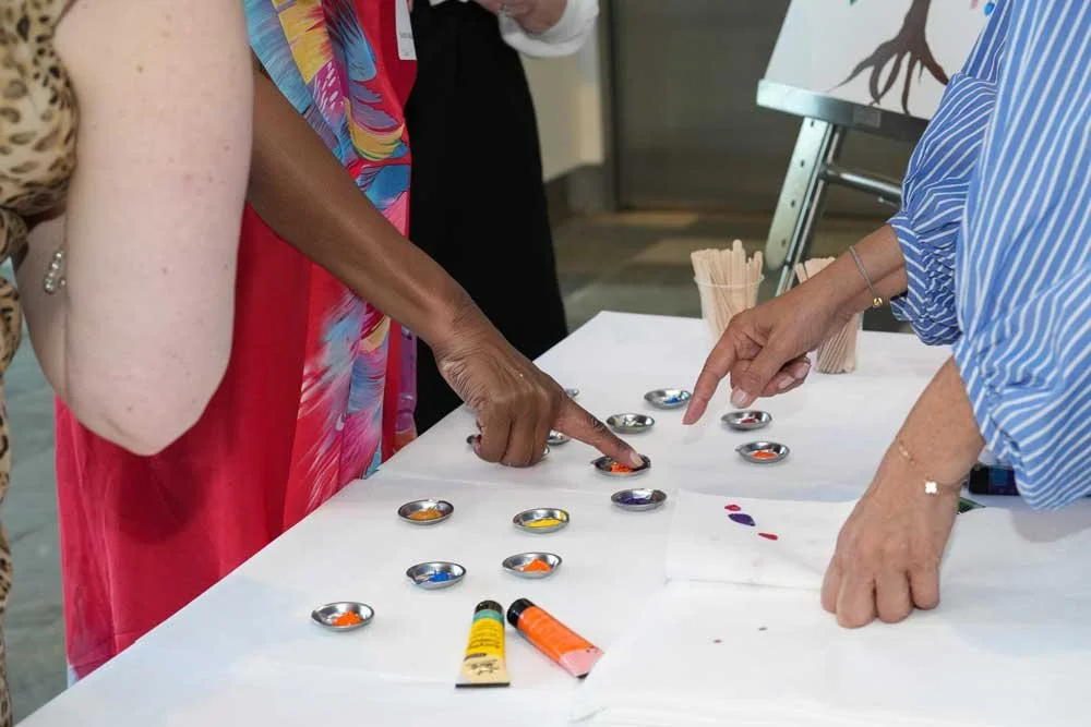 Two women pointing at colors in small containers on a table, with art supplies nearby.