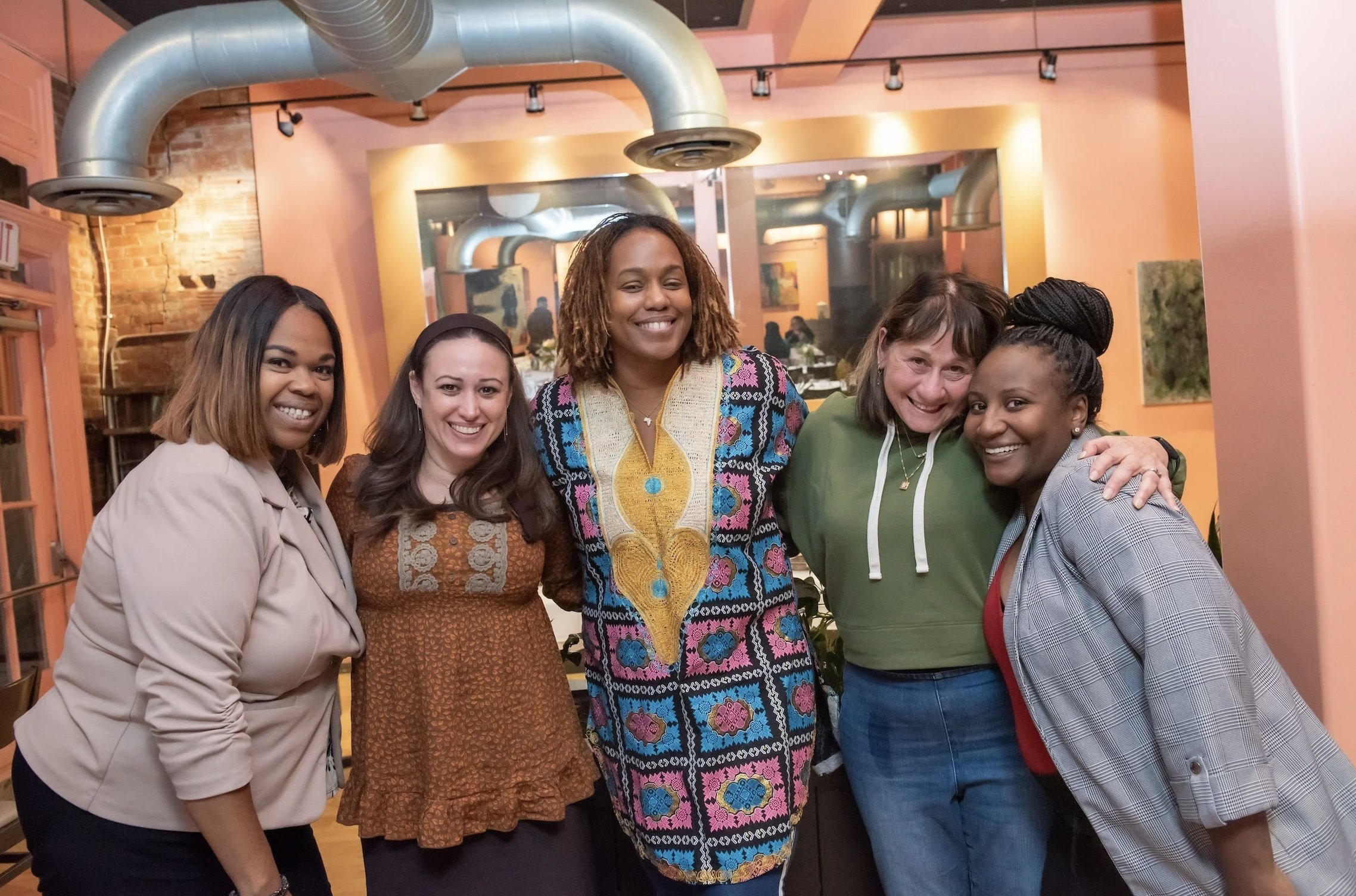 Five women smiling and posing for a photo indoors, with industrial-style ductwork and artwork on the walls in the background.