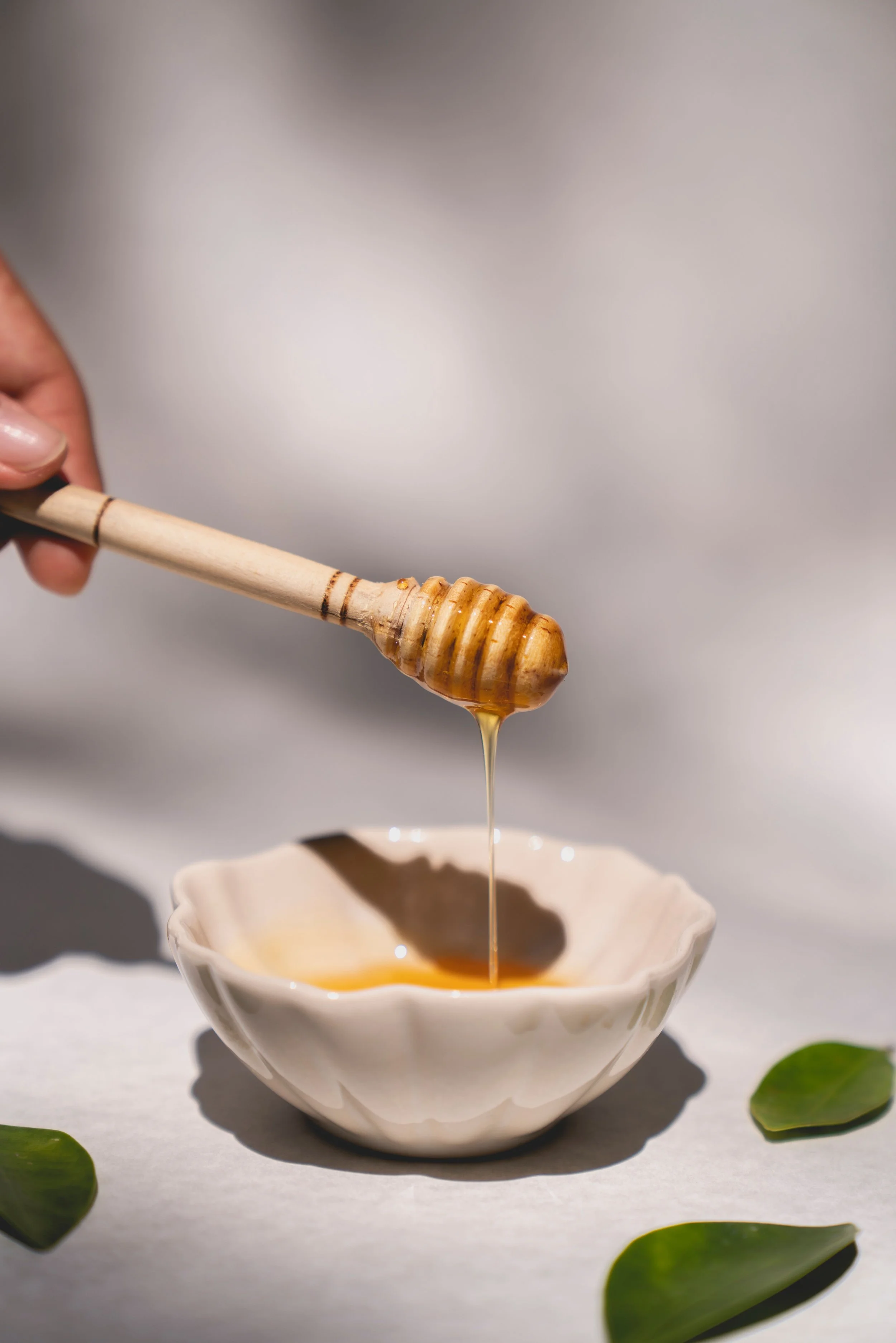 A hand holding a honey dipper drizzling honey into a ceramic bowl placed on a white surface surrounded by green leaves.