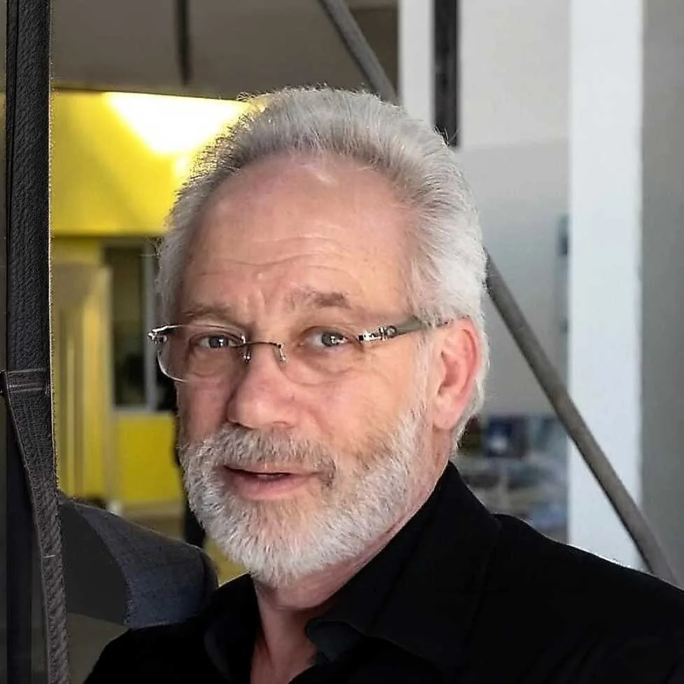 A smiling older man with white hair and beard, wearing glasses and a black shirt, in an indoor setting.