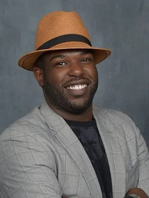 A smiling man wearing a tan fedora hat, gray plaid blazer, and black shirt, posing against a plain dark gray background.