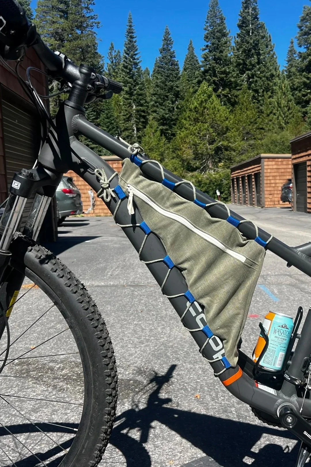 A mountain bike with a frame bag parked in a lot surrounded by tall pine trees.