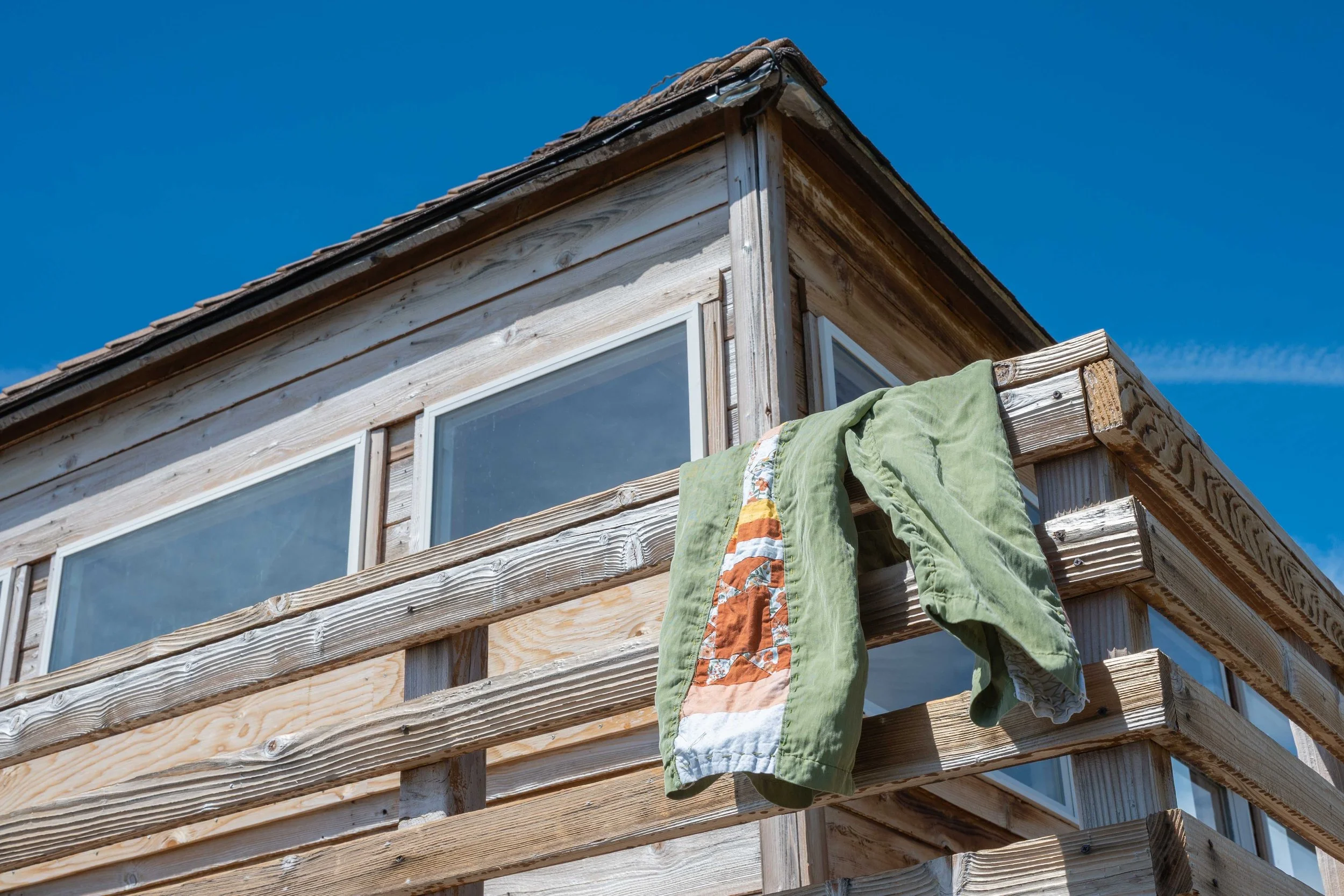 A fire-tower lookout with large windows and an unfinished exterior. Clothes, including green pants with a quilted strip, are hanging on a railing outside.