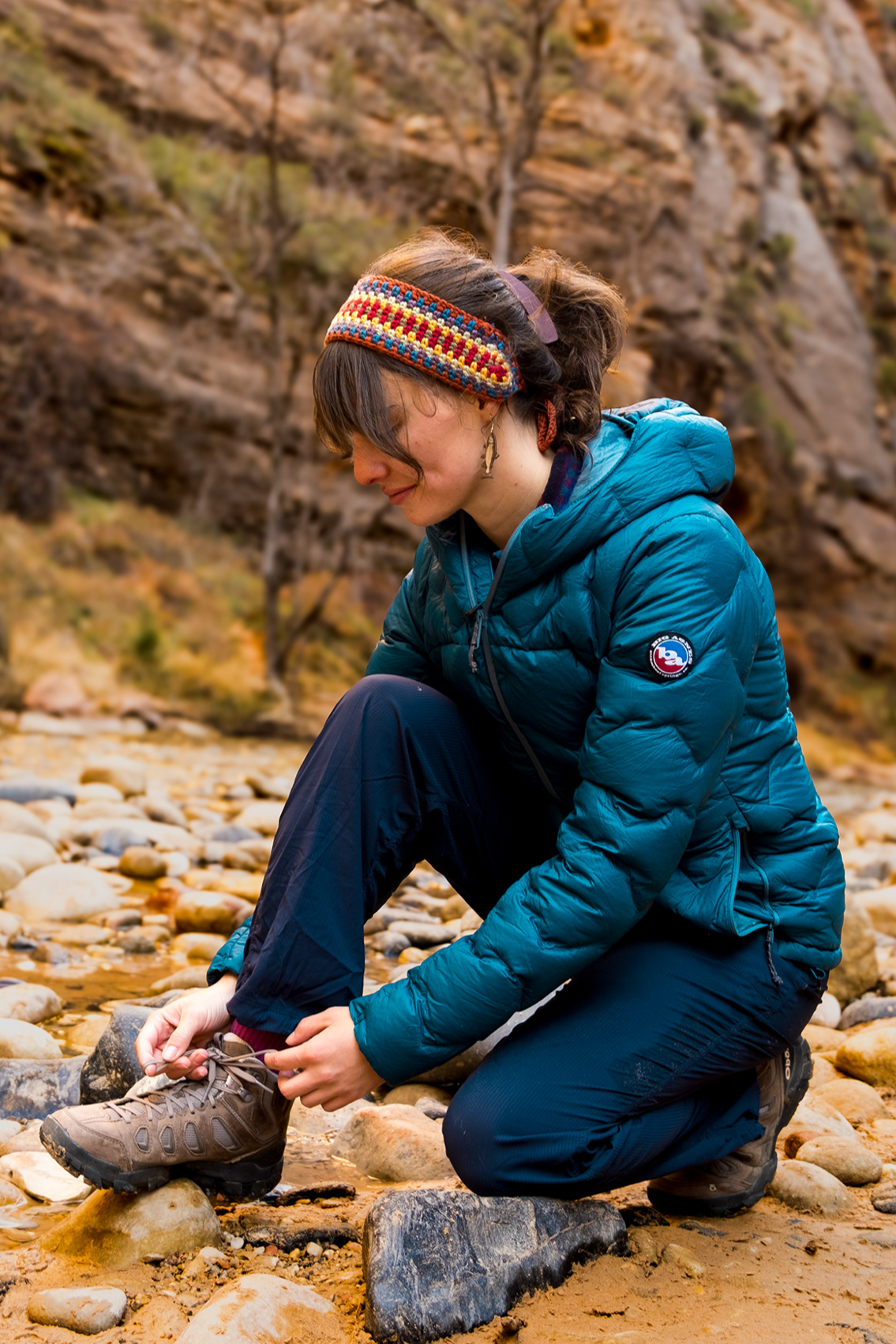 Woman kneeling on rocky riverbank, tying her hiking boot, wearing a blue puffer jacket, colorful headband, and outdoor gear, with rocky cliffs and sparse trees in the background.